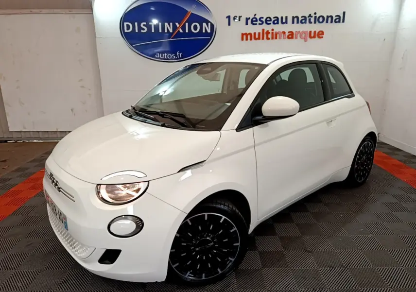 Fiat 500 électrique blanc vue 3/4 avant droit en intérieur avec jantes noires et phares allumés.