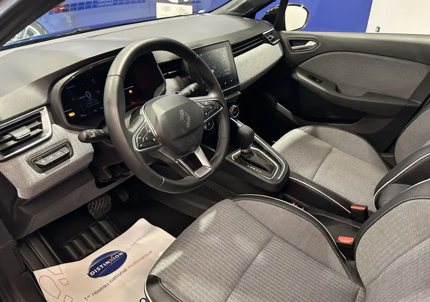Vue intérieure côté conducteur de la Renault Clio V hybride, avec tableau de bord numérique et sellerie tissu gris.
