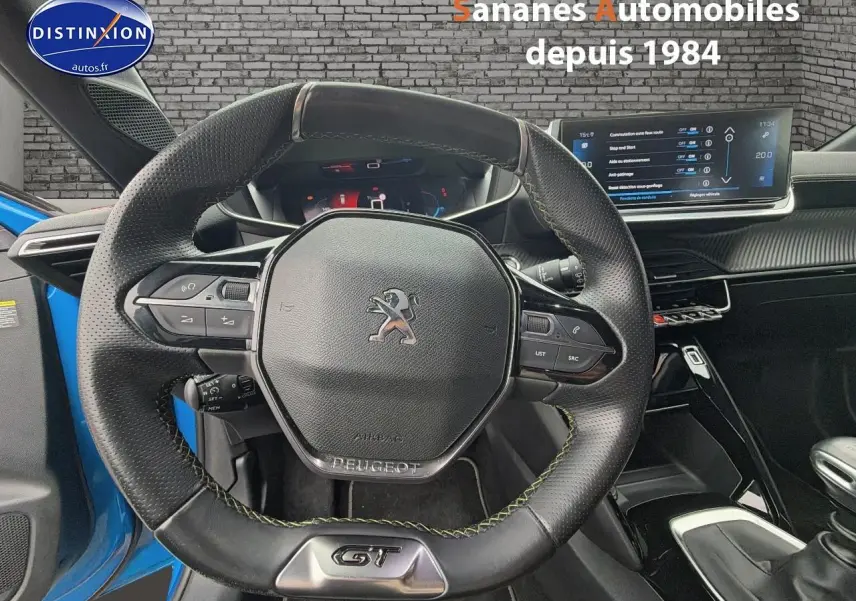 Vue centrée sur le volant cuir noir GT avec coutures vertes, tableau de bord numérique et écran tactile du Peugeot 208 bleu clair.
