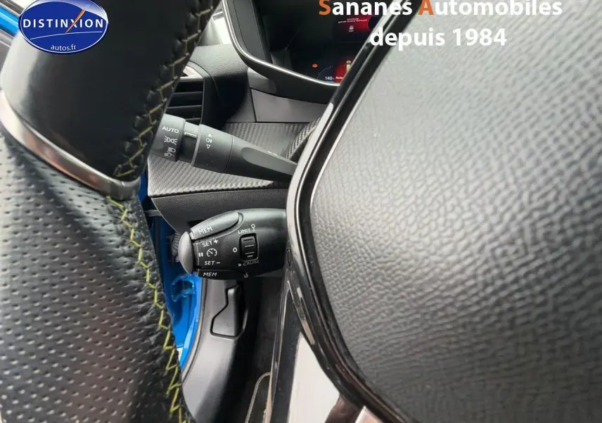 Vue rapprochée du volant cuir noir avec surpiqûres vertes et commandes, tableau de bord partiel d'une Peugeot 208 bleu clair 2020.