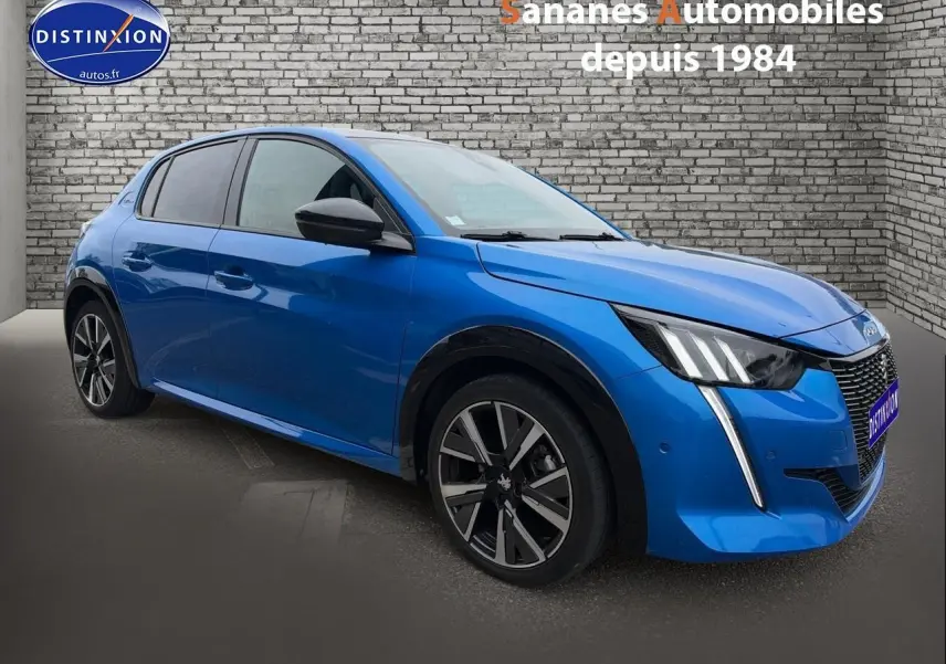 Peugeot 208 bleu clair en 3/4 avant droit, avec jantes alliage et feux LED distinctifs, sur fond mur en briques.