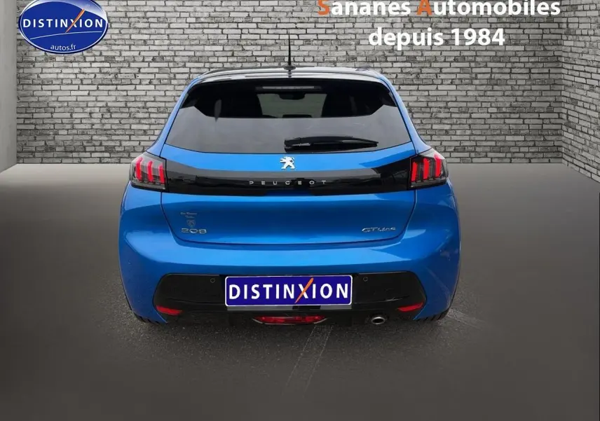 Vue arrière d'une Peugeot 208 GT Line bleu clair avec feux arrière LED et lunette teintée, sur fond gris et mur en briques.
