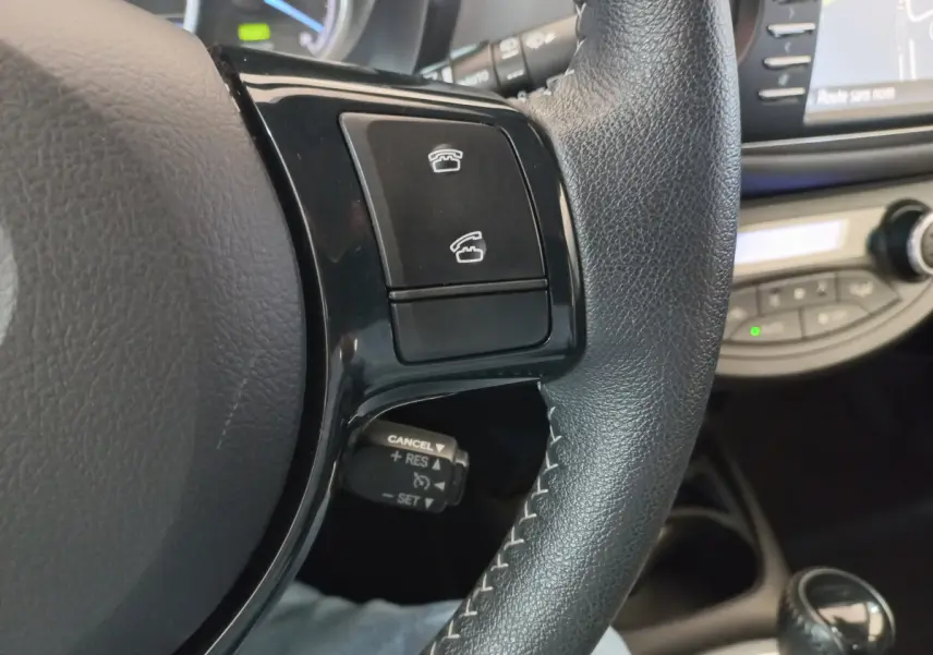 Gros plan sur le volant noir en cuir de la Toyota Yaris blanche, avec commandes téléphoniques et régulateur de vitesse visible.