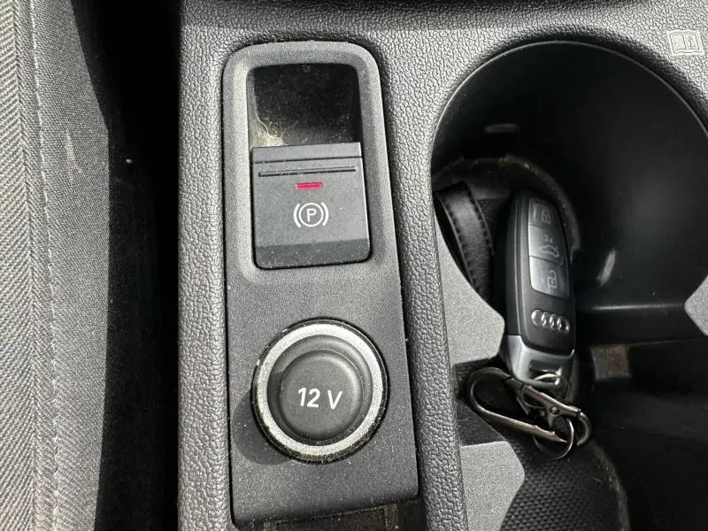 Gros plan sur la console centrale noire de l'Audi A3 Sportback 35 TFSI, avec bouton frein à main électrique et prise 12V, clé posée à côté.