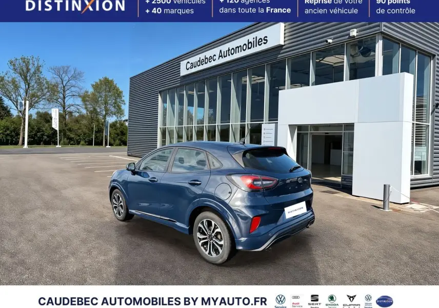 Ford Puma bleu en 3/4 arrière droit, avec jantes alliage 17 pouces et feux arrière LED visibles, devant un showroom.