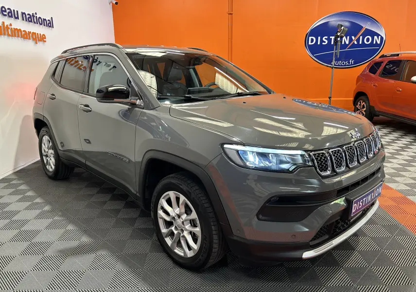 Jeep Compass gris en 3/4 avant droit avec phares allumés, présenté en intérieur sur sol damier noir et gris.