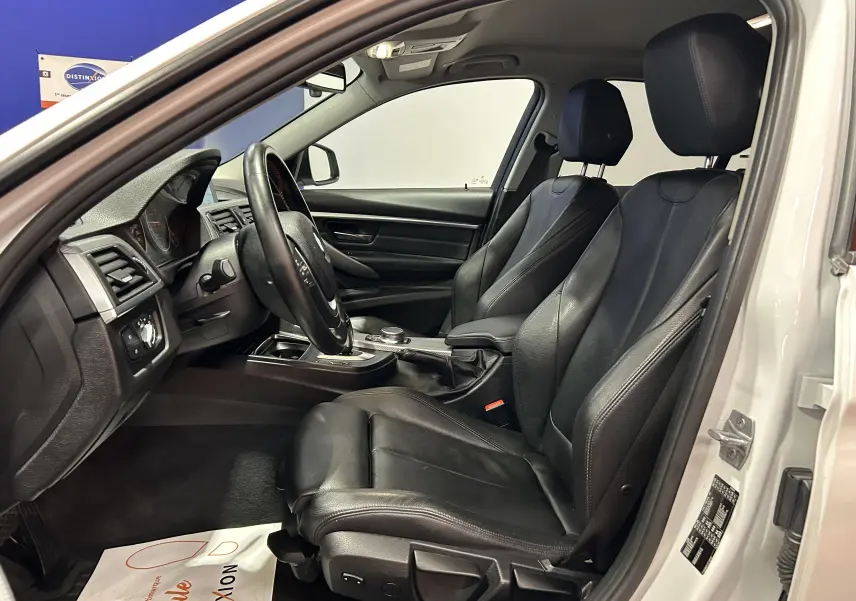 Vue intérieure côté conducteur de la BMW Série 3 320i blanche avec sièges cuir noirs et volant multifonction.