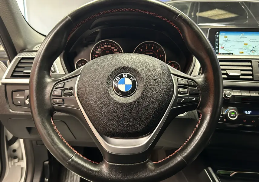 Vue rapprochée du volant cuir noir avec coutures rouges d'une BMW Série 3 320i 2018, tableau de bord et écran GPS visibles.