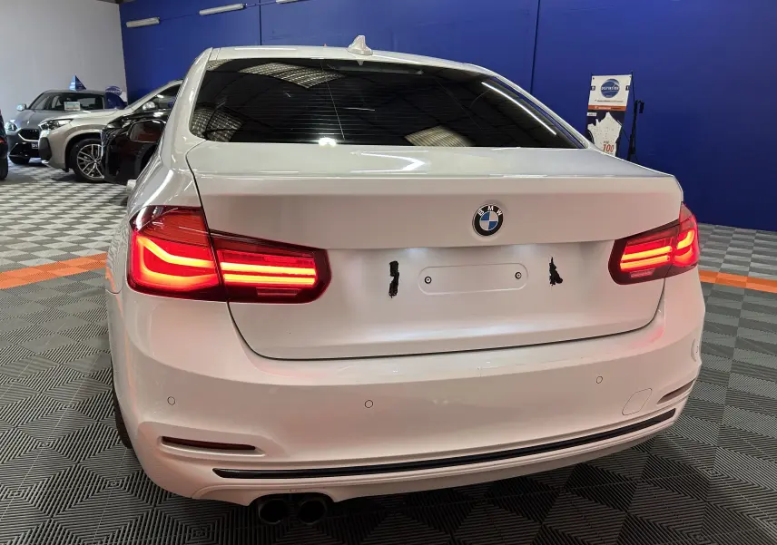 Vue arrière d'une BMW Série 3 320i blanche avec feux allumés dans un showroom intérieur.