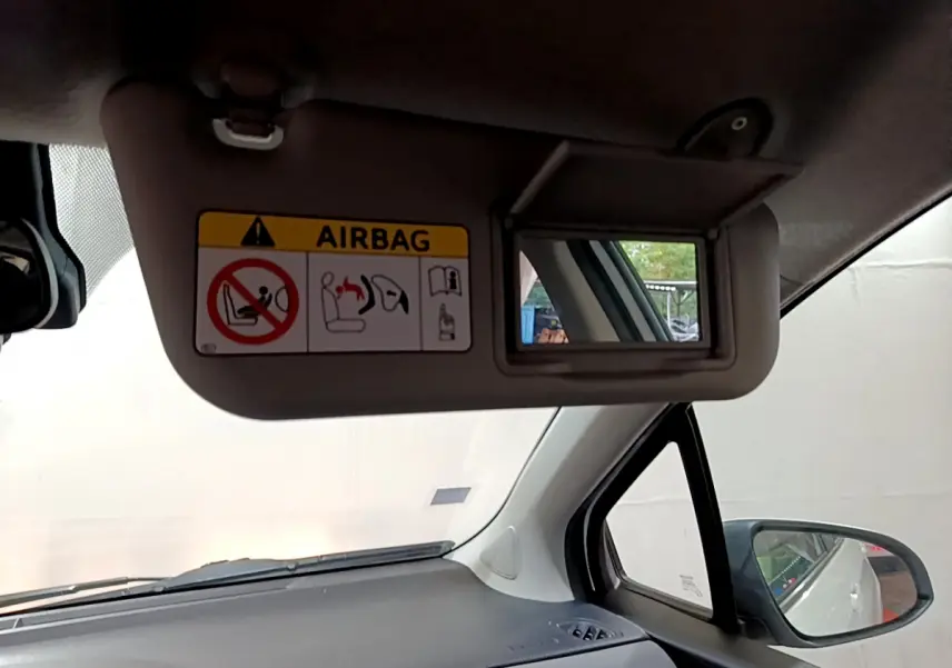 Miroir de pare-soleil côté conducteur avec avertissement airbag, tableau de bord gris et rétroviseur extérieur visible sur Toyota Yaris blanche.