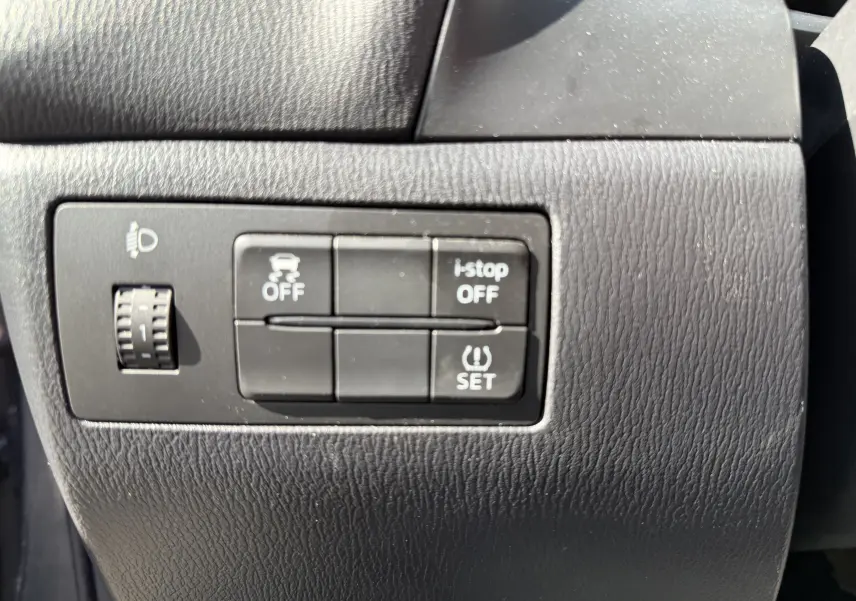 Gros plan sur les boutons de commande noirs du tableau de bord de la Mazda3 gris, avec réglage des phares et fonctions i-stop et contrôle de stabilité.
