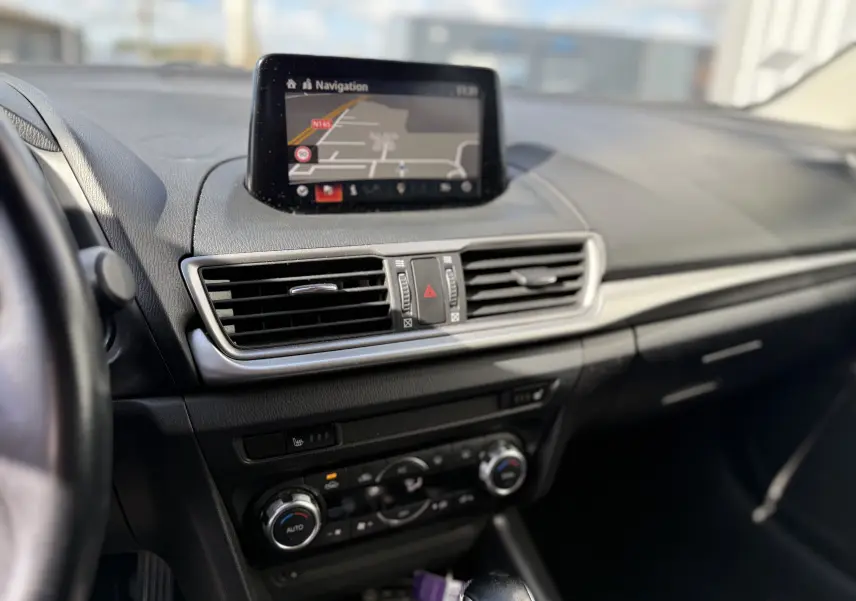Vue intérieure centrée sur le tableau de bord de la Mazda3 2017, écran navigation et commandes climatisation visibles.