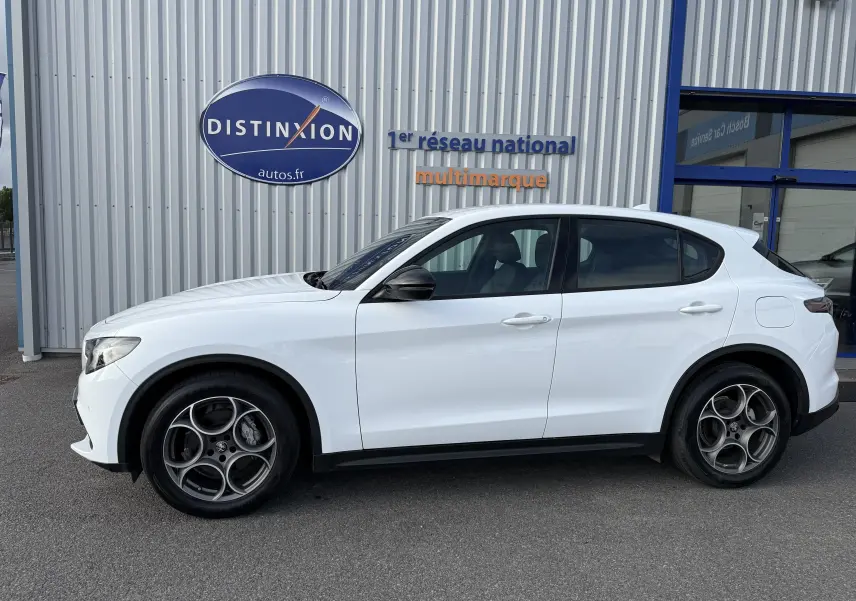 Profil côté droit de l'Alfa Romeo Stelvio blanc Alfa 2023, avec jantes spécifiques et rétroviseurs noirs, devant un bâtiment métallique.