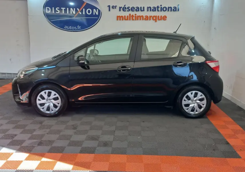 Profil côté gauche d'une Toyota Yaris noire 2018 hybride, avec jantes acier et enjoliveurs, en intérieur showroom.