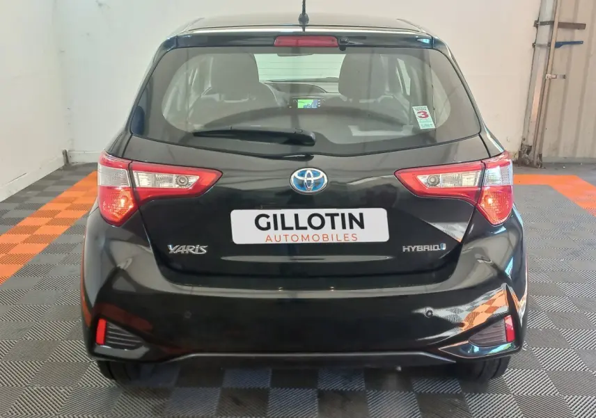 Vue arrière d'une Toyota Yaris hybride noire avec feux arrière allumés et logo visible sur un sol à damiers noir et orange.