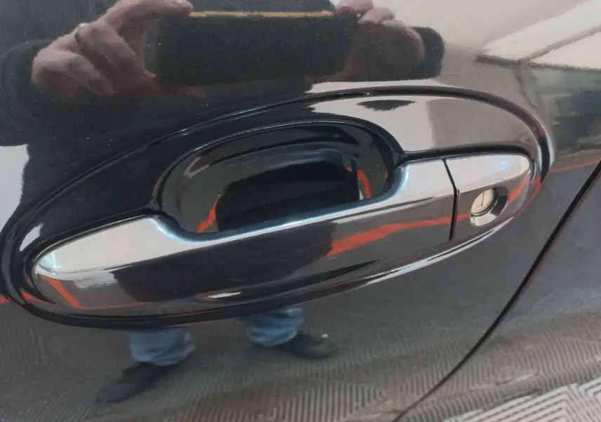 Poignée de porte avant droite chromée sur Toyota Yaris noire, reflet de la personne prenant la photo visible.