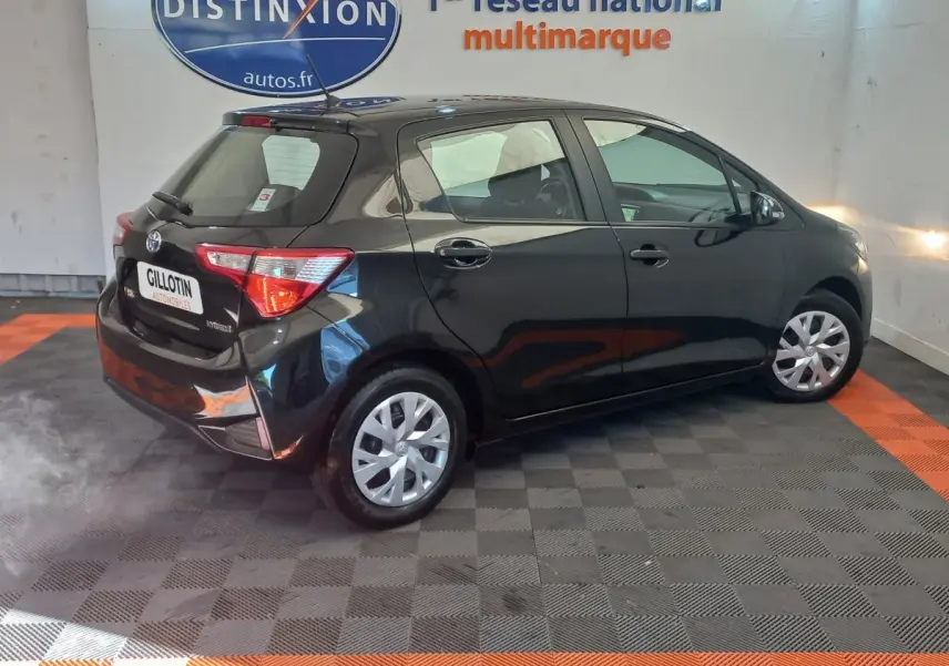 Toyota Yaris noire vue de trois quarts arrière côté droit dans un showroom avec sol gris et orange.