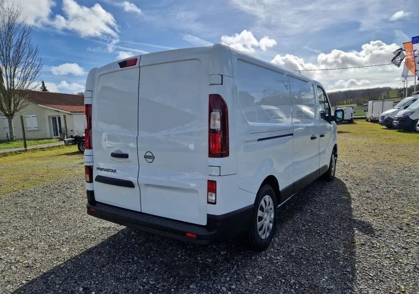Vue 3/4 arrière droite d’un utilitaire Nissan Primastar blanc stationné sur un terrain gravillonné par temps ensoleillé.