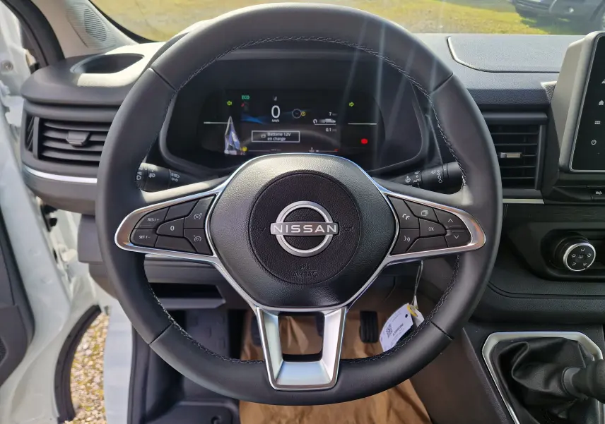 Vue intérieure centrée sur le volant noir du Nissan Primastar blanc avec tableau de bord numérique et levier de vitesse manuel.
