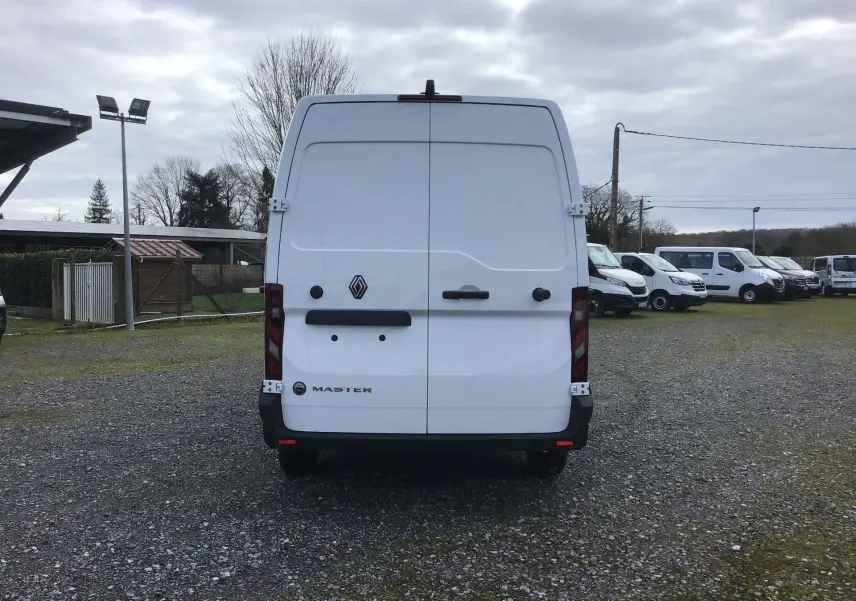 Vue arrière d’un utilitaire Renault Master blanc 2025, portes battantes fermées, sur un terrain extérieur avec plusieurs autres véhicules.