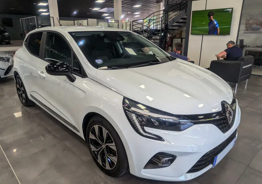 Renault Clio V TCe 100 GPL blanc glacier vue 3/4 avant droit en intérieur avec jantes noires bicolores.