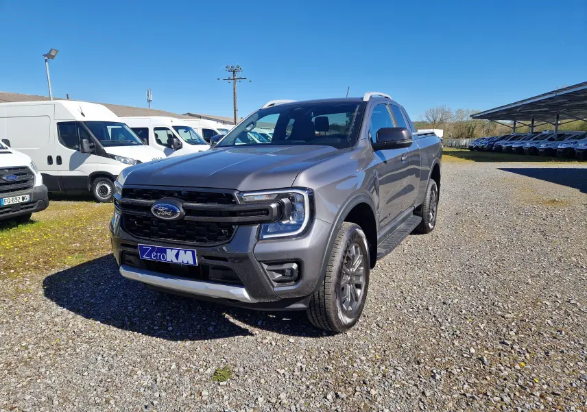 Ford Ranger Super Cabine gris carbone vu en 3/4 avant droit sur un sol gravillonné avec plusieurs fourgons blancs en arrière-plan.