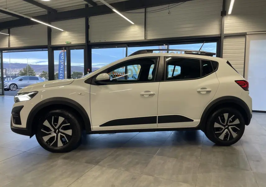 Vue de profil côté gauche d'une Dacia Sandero Stepway 2025 blanc glacier avec roue de secours visible en intérieur showroom.