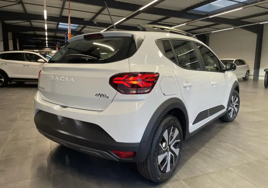 Vue 3/4 arrière droite d'une Dacia Sandero Stepway Expression 2025 blanc glacier avec roue de secours en intérieur showroom.