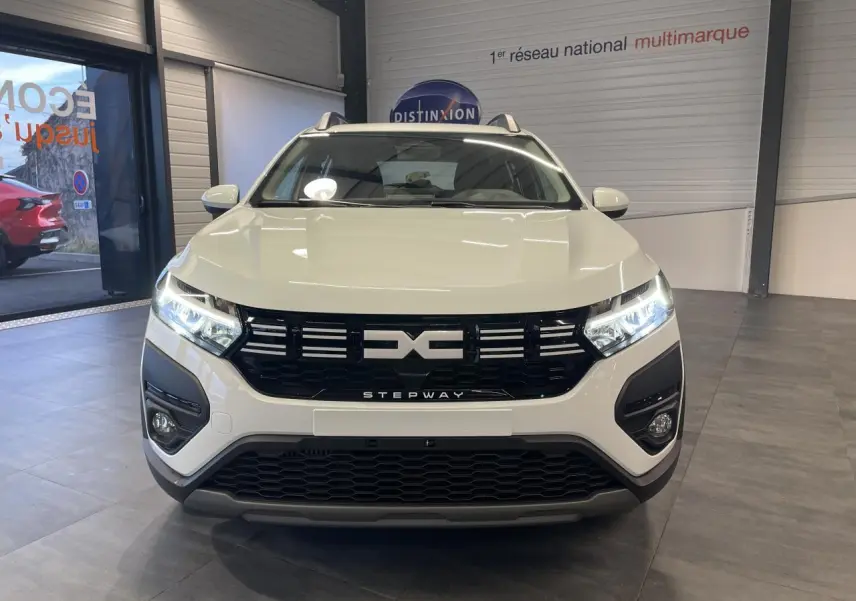 Vue avant d'une Dacia Sandero Stepway Expression 2025 blanc glacier avec calandre noire et phares allumés en intérieur showroom.