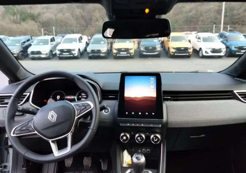 Vue intérieure avant de la Renault Clio V TCe 90 Techno Plus 2025, tableau de bord gris avec écran tactile vertical et volant multifonctions.