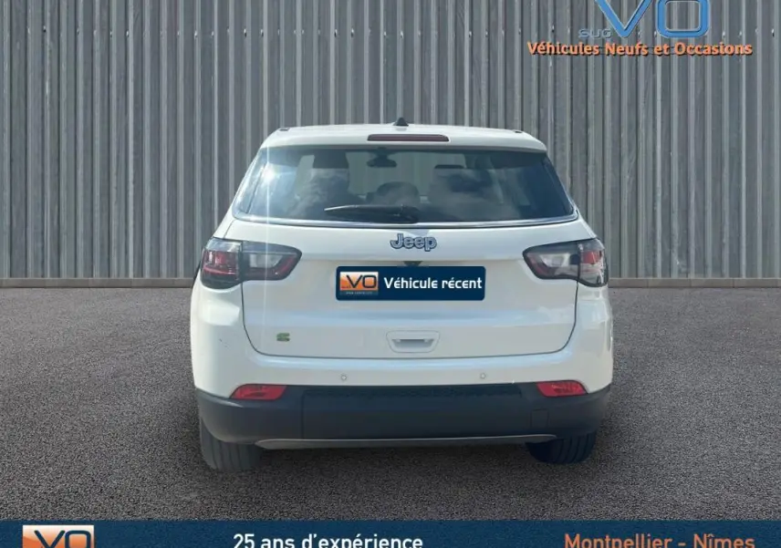 Vue arrière d'un Jeep Compass blanc 2024 avec roue de secours et jantes 17 pouces, stationné devant un mur métallique.