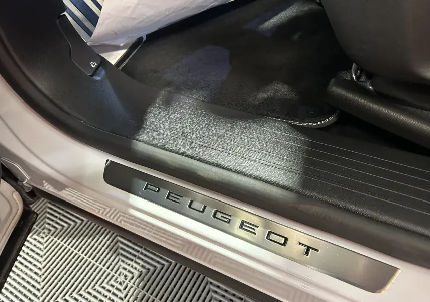 Gros plan sur la plaque de seuil de porte Peugeot sur un véhicule blanc, intérieur avec tapis noir visible.