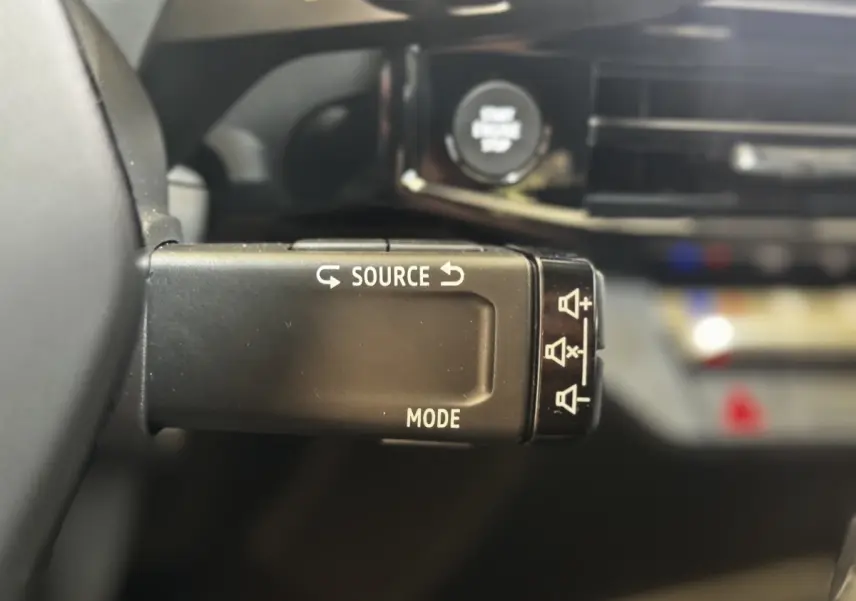 Gros plan sur la commande au volant de la Renault Clio 2026, bouton source et mode, intérieur noir flouté en arrière-plan.