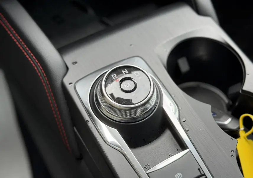 Gros plan sur le sélecteur rotatif de boîte automatique et accoudoir noir avec surpiqûres rouges dans un Ford Kuga noir Agathe.