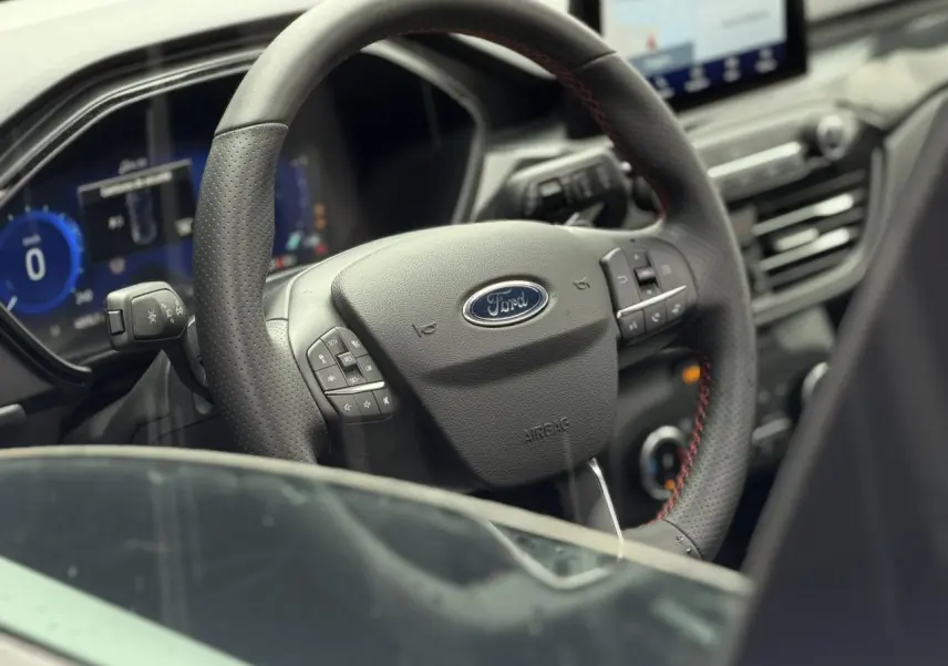 Vue rapprochée du volant noir perforé avec surpiqûres rouges du Ford Kuga 2023, tableau de bord et écran tactile visibles.