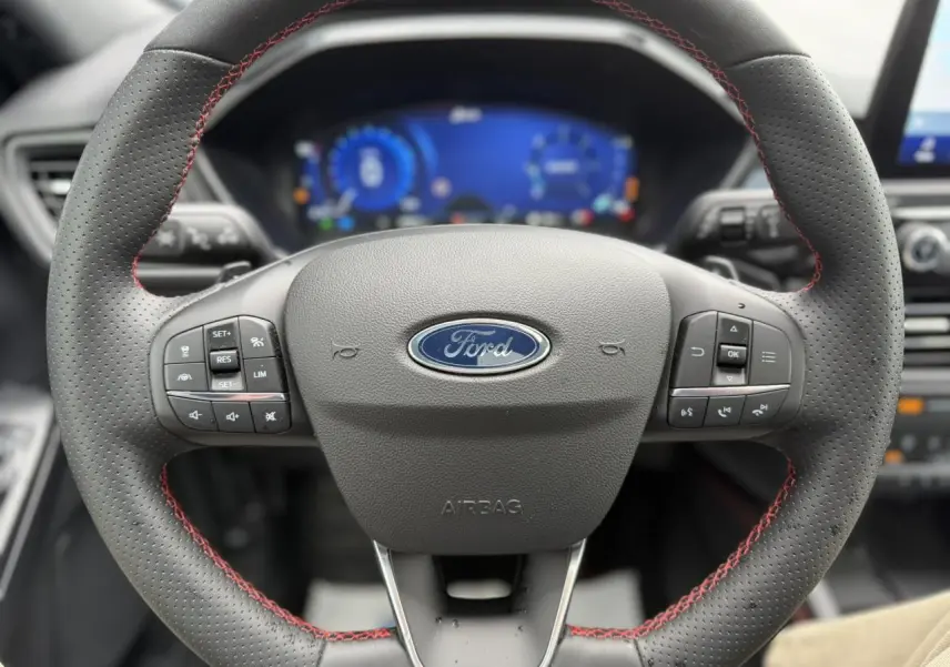 Vue rapprochée du volant perforé noir avec surpiqûres rouges du Ford Kuga 2023, tableau de bord numérique en arrière-plan.