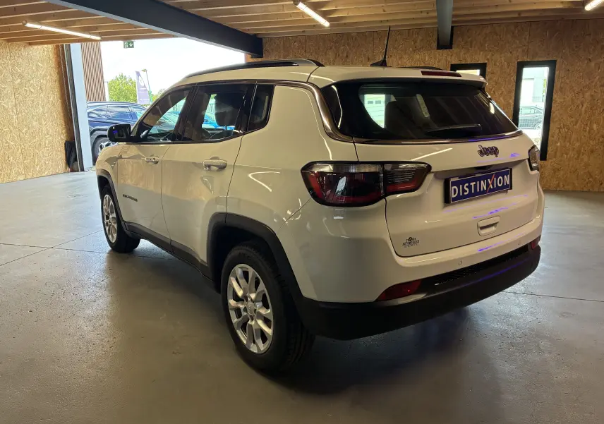 Jeep Compass ivoire vu en 3/4 arrière droit dans un garage, avec barres de toit et jantes alu visibles.