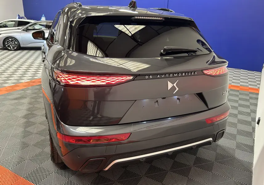 Vue arrière 3/4 d'un DS7 gris titane avec feux LED distinctifs et logo DS Automobiles sur fond intérieur showroom.