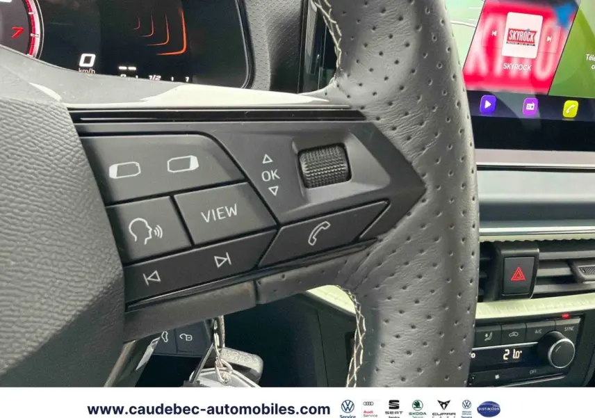 Gros plan sur le volant en cuir perforé de la SEAT Arona blanche, avec commandes multifonctions et écran tactile visible.