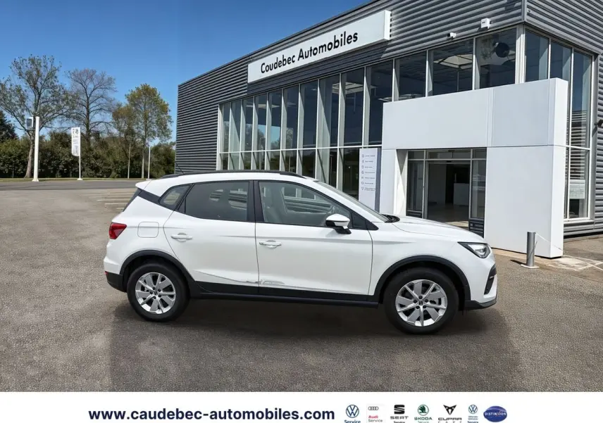 Vue de profil côté gauche d'un SEAT Arona blanc garé devant un bâtiment moderne de concession automobile.
