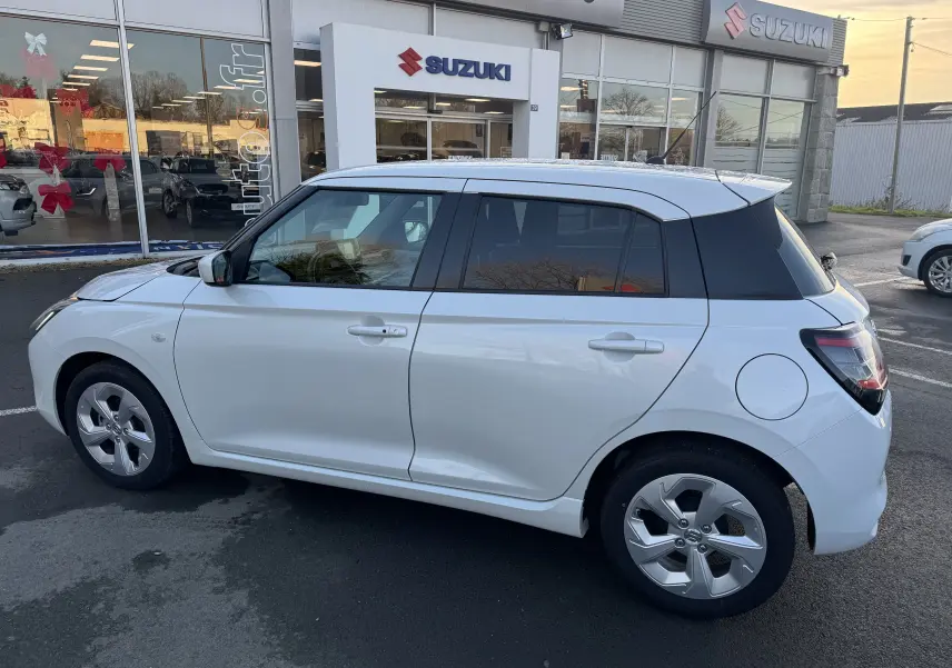Profil côté droit d'une Suzuki Swift 1.2 Privilège Hybrid blanche, compacte et moderne, stationnée devant un concessionnaire.