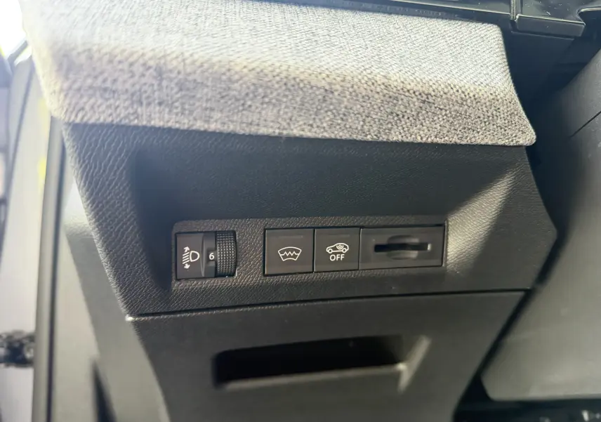 Détail intérieur côté gauche du tableau de bord gris artense de la Peugeot 1007 Hybrid avec commandes de réglage des phares et aides à la conduite.
