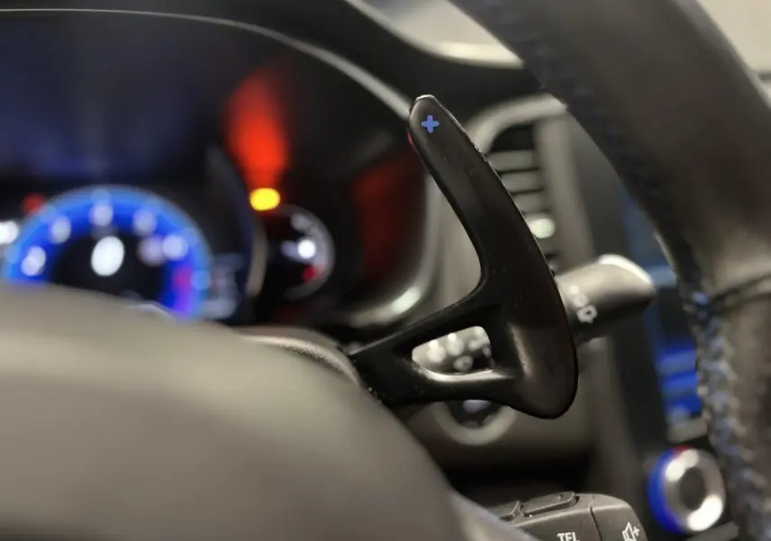 Gros plan sur la palette de changement de vitesse + au volant cuir perforé de la Renault Mégane GT bleue, tableau de bord flou en arrière-plan.