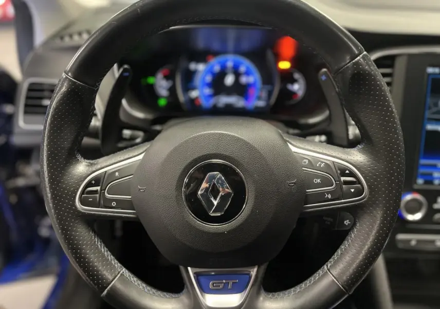 Gros plan sur le volant cuir perforé noir avec logo Renault et badge GT, tableau de bord numérique en arrière-plan.