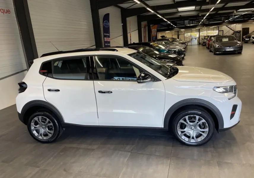 Vue de profil côté gauche d'une Citroën C3 blanche avec barres de toit noir brillant en showroom.