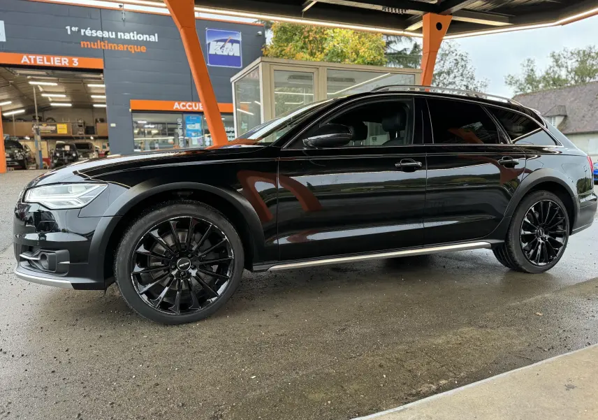 Audi A6 Avant Allroad noire vue de profil côté gauche, avec jantes noires et barres de toit visibles.