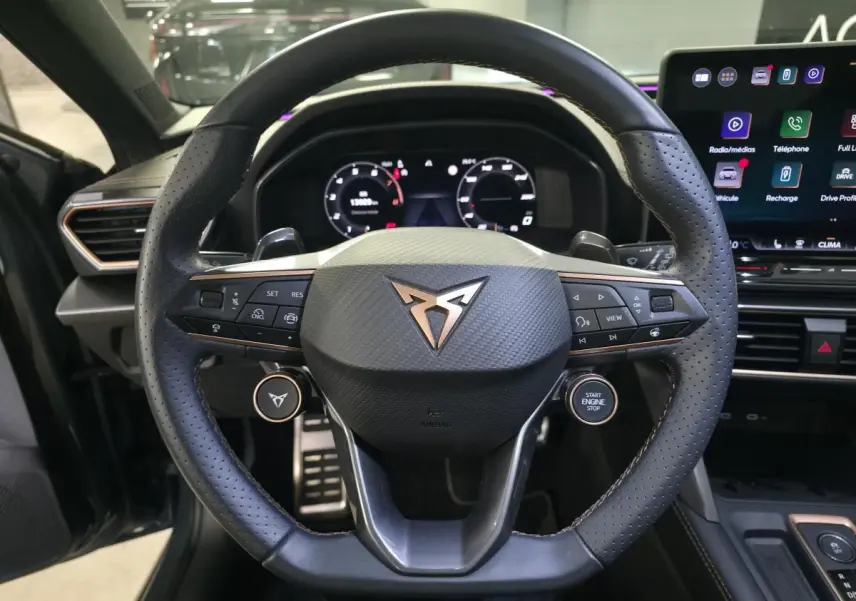 Vue rapprochée du volant cuir noir perforé avec logo CUPRA cuivré et commandes intégrées, tableau de bord digital en arrière-plan.