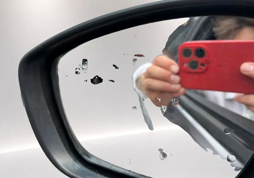 Rétroviseur extérieur gauche d’un Opel Mokka gris 2025 avec traces d’eau et reflet d’une main tenant un smartphone rouge.