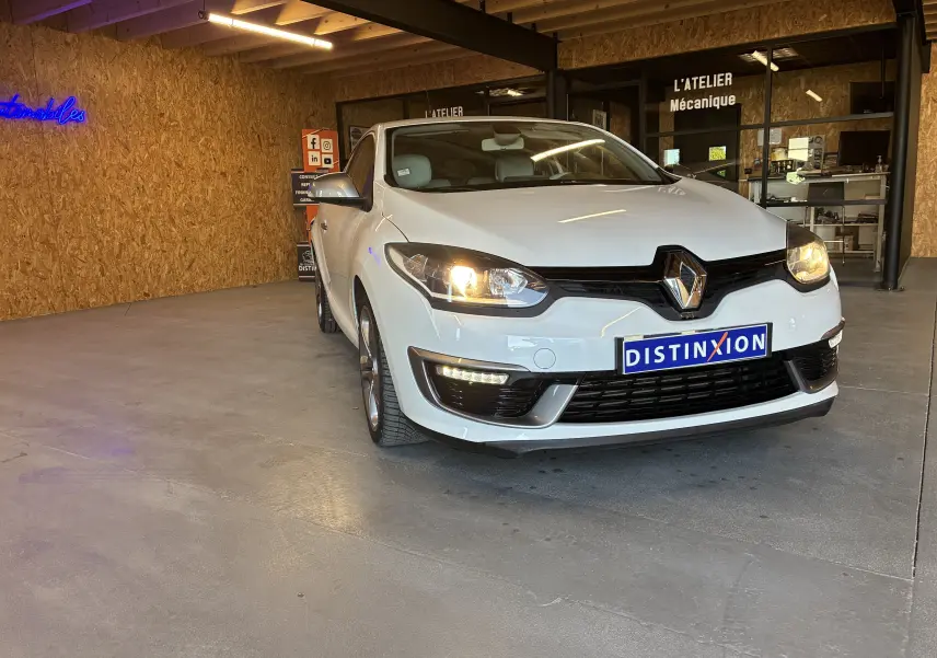 Renault Mégane Coupé blanc vue 3/4 avant droit avec phares allumés dans un garage intérieur.