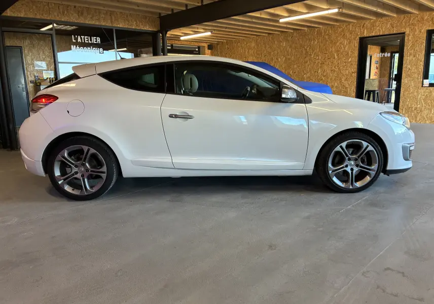 Profil côté gauche d'une Renault Mégane Coupé blanche de 2014, avec jantes alliage et rétroviseurs argentés dans un garage.
